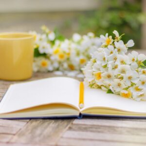 open notebook , yellow cup and white flowers on wooden table in garden