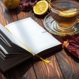 Open notebook with blank pages on a wooden table, accompanied by a hot cup of lemon tea, a warm sweater, and vibrant autumn leaves, creating a cozy atmosphere in morning light