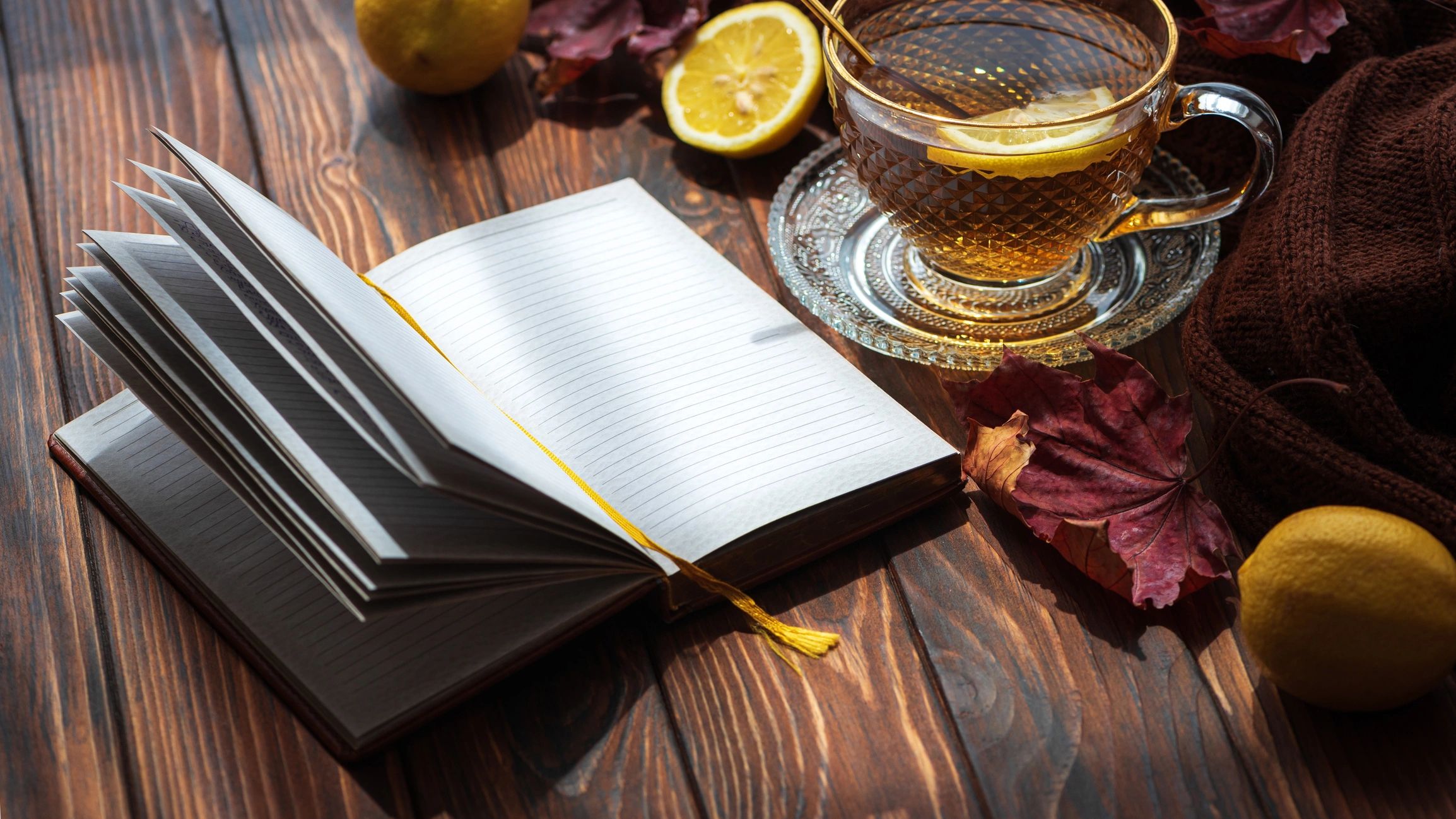 Open notebook with blank pages on a wooden table, accompanied by a hot cup of lemon tea, a warm sweater, and vibrant autumn leaves, creating a cozy atmosphere in morning light
